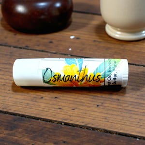 May include: White lip balm tube with the word "Osmanthus" in script, and a floral design with yellow and blue flowers. The tube also lists ingredients like coconut oil and beeswax.