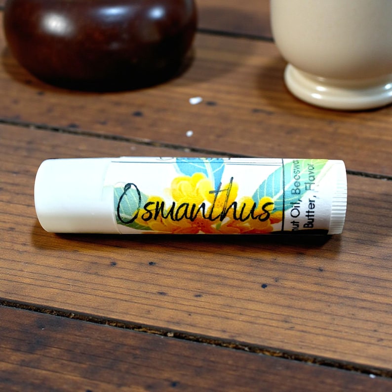 May include: White lip balm tube with the word "Osmanthus" in script, and a floral design with yellow and blue flowers. The tube also lists ingredients like coconut oil and beeswax.