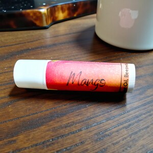 May include: A lip balm tube with a white cap and a label featuring a gradient of red, orange, and yellow. The word "Mango" is written in black script on the label. The label also includes the words "Oil, Beeswax".
