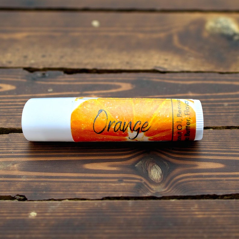 May include: A white lip balm tube with an orange label featuring the word "Orange" in cursive. The label also includes a graphic of an orange slice. The lip balm is resting on a wooden surface.