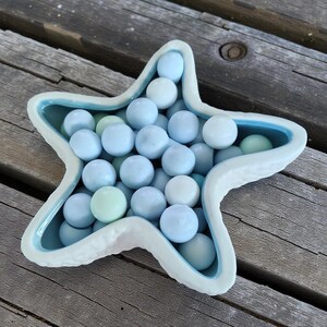 May include: A ceramic starfish-shaped bowl filled with light blue marbles. The bowl has a textured, white exterior and a light blue interior. The marbles vary in shades of blue and green. The bowl is placed on a weathered wooden surface.
