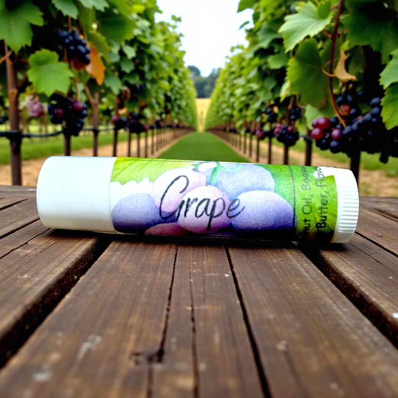 May include: A white lip balm tube with a label featuring a watercolor illustration of purple grapes and the word "Grape". The tube is lying on a wooden surface, with a vineyard in the background.