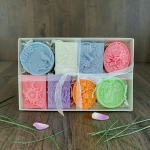 May include: A gift box filled with eight decorative soaps in various shapes and colors. The soaps feature floral and bird designs, including a blue round soap with a bird and flowers, and a pink oval soap with a peony. The soaps are tied with a white ribbon.