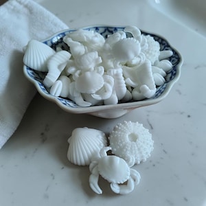 May include: A small, blue and white patterned bowl filled with white seashell and crab-shaped decorative items. Additional white seashell, crab, and sea urchin-shaped items are arranged around the bowl. The items are made of a solid, opaque material.