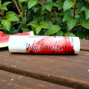 May include: A white lip balm tube with a watermelon design and the word "Watermelon" in script. Slices of watermelon are in the background on a wooden surface, with green foliage in the background.