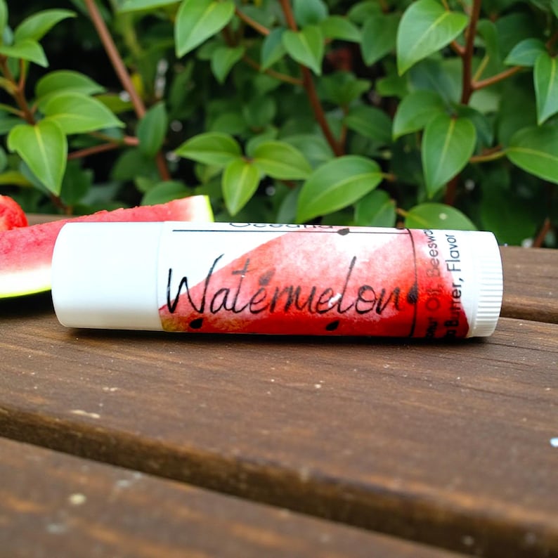 May include: A white lip balm tube with a watermelon design and the word "Watermelon" in script. Slices of watermelon are in the background on a wooden surface, with green foliage in the background.