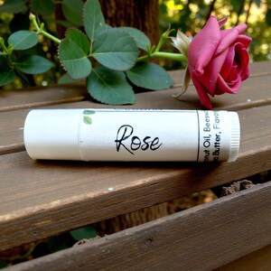 May include: A white lip balm tube with the word "Rose" in black script. A pink rose and green leaves are in the background. The lip balm contains coconut oil, beeswax, and cocoa butter.