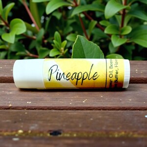 May include: A lip balm tube with a white cap and a yellow label. The label reads "Pineapple" in a handwritten font. The background is a wooden surface with green foliage.