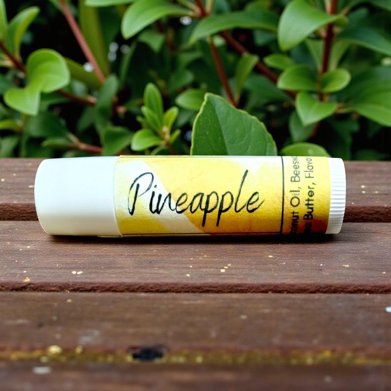 May include: A lip balm tube with a white cap and a yellow label. The label reads "Pineapple" in a handwritten font. The background is a wooden surface with green foliage.