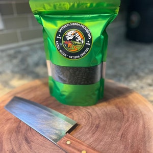 May include: A green bag of coffee beans with a label that says "Saffron Sierra Provisions Est'd 2020 Small Batch Artisan Crafted" and a mountain scene. The bag is sitting on a wooden cutting board with a silver knife.