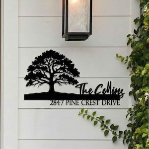 Peut inclure: Panneau d'adresse en métal noir avec une silhouette d'arbre, le nom de famille "The Collins" et l'adresse "2847 Pine Crest Drive". Le panneau est monté sur un mur blanc à côté d'une porte en bois et d'un luminaire extérieur noir.