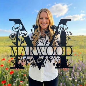 Może przedstawiać: Czarny metalowy monogram kwiatowy z wyciętym imieniem "MARWOOD". Litera "M" jest ozdobiona kwiatowymi wzorami. Osoba trzyma monogram na tle pola kwiatów.