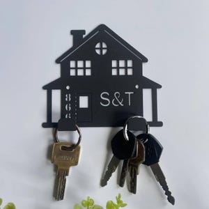 May include: Black metal house-shaped key holder with the letters "S&T" and the numbers "863" cut out. The key holder has a roof, windows, and a door. Several keys hang from the bottom.
