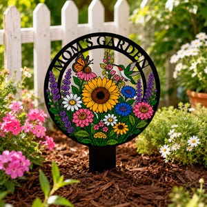 May include: A circular black metal garden stake featuring a colorful floral design with a large yellow sunflower, pink and blue flowers, a butterfly, and a hummingbird. The text MOM'S GARDEN is cut out along the top edge of the circular frame.