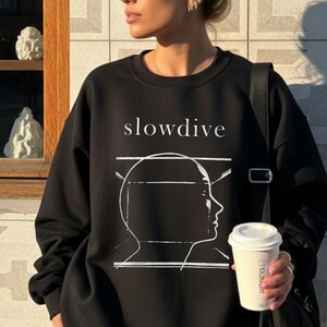 May include: Black sweatshirt and shorts set. The sweatshirt features the word "slowdive" above a white graphic of a person's profile. The person is wearing sunglasses and holding a coffee cup. The background is a tiled wall and a window.
