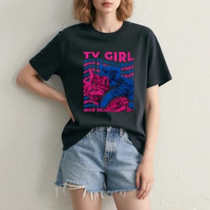 以下が含まれることがあります： ピンクと青で「TV GIRL」の文字が入った黒いTシャツ。シャツには「WHO REALLY CARES」という言葉が繰り返されるグラフィックデザインが施されています。Tシャツはデニムショーツと組み合わせています。