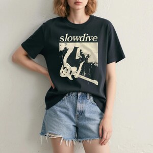 May include: Black t-shirt with the band name "slowdive" in white block letters above a white and black graphic of a person playing a guitar. The model is wearing denim shorts.