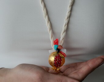 May include: A white rope necklace with a gold pomegranate pendant. The pendant is decorated with red, orange, and turquoise stones.