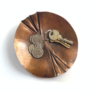 May include: A handcrafted, copper-colored metal leaf-shaped dish. The dish contains several quarters and a set of keys. The dish is designed with textured details and is ideal for holding small items like keys and coins.