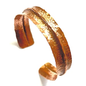 May include: A copper cuff bracelet featuring a distinct hammered texture across its surface. The metal has a warm, reddish-brown metallic finish with a curved, open-ended design that creates a rippled, wave-like appearance along the outer edge.