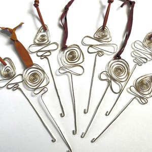 Puede incluir: Colección de adornos artesanales de alambre metálico con diseños geométricos y en espiral. Cada adorno tiene un tallo largo y delgado y está rematado con un lazo de cuero o tela en tonos marrón y burdeos.