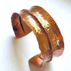 May include: A wide, open-ended cuff bracelet crafted from polished copper. The metal surface features a distinct hammered texture with an irregular, wavy edge profile that creates a rippled appearance along the entire length of the band.