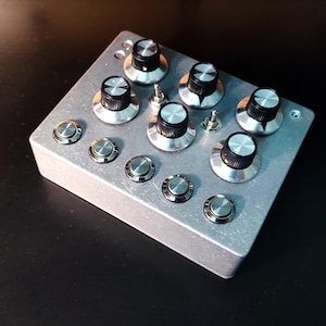May include: A silver electronic device featuring multiple knobs and buttons. The rectangular unit has a brushed metal finish. It has eight large knobs with black tops and silver bases, plus several smaller silver buttons.