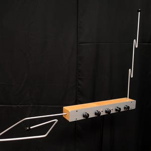 Puede incluir: Un instrumento musical electrónico theremin con una base rectangular de madera y un panel de control de metal plateado. Incluye cinco perillas circulares negras, dos interruptores de palanca, una antena vertical en zigzag y una antena de bucle.