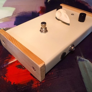 Puede incluir: Un pedal de efectos blanco con una carcasa de madera. El pedal tiene un solo pedal, una perilla y una entrada jack.