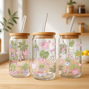 May include: Three clear glass tumblers with wooden lids and glass straws. Each tumbler features a different design of potted plants, pink bows, and hearts. The tumblers are on a wooden surface.