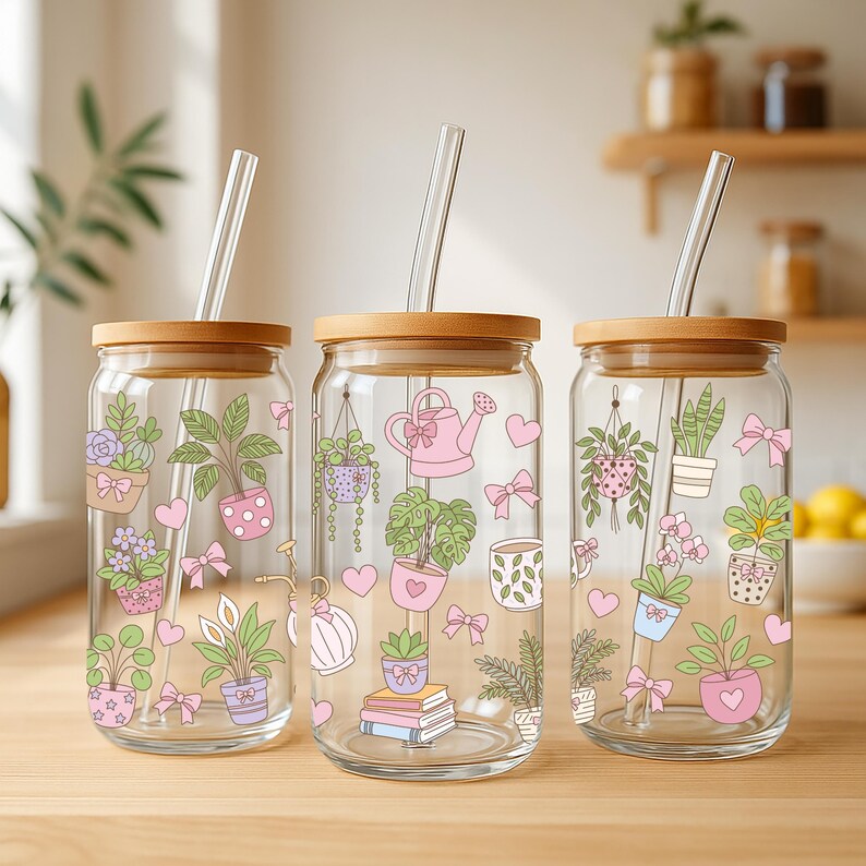 May include: Three clear glass tumblers with wooden lids and glass straws. Each tumbler features a different design of potted plants, pink bows, and hearts. The tumblers are on a wooden surface.