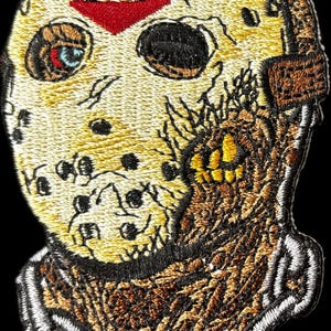 May include: Embroidered patch featuring a distressed hockey mask with a red chevron. The mask is primarily tan with brown and black details, and a stitched white border. The patch has a horror theme.