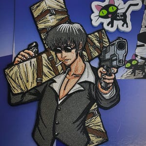 Puede incluir: Parche bordado con un hombre en traje sosteniendo una pistola, con una cruz detrás de él. La cruz está envuelta en vendas y tiene un cigarrillo colgando. El hombre lleva gafas de sol y tiene una expresión seria. El parche es en blanco y negro.