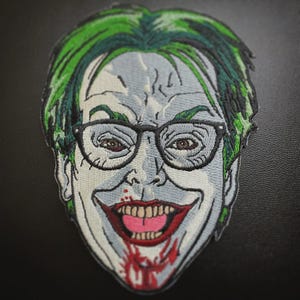 May include: Embroidered patch featuring a stylized face with green hair, glasses, and a wide, bloody grin. The patch has a gray and white color scheme with brown eyes and red accents. The design is a portrait of a comic book villain.