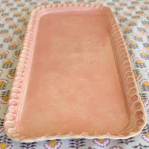 Puede incluir: Una bandeja rectangular de color rosa claro con una superficie texturizada. La bandeja tiene un borde festoneado con un patrón repetitivo de pequeños círculos en relieve. La bandeja se muestra sobre un mantel con estampado floral.
