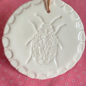 Peut inclure: Ornement rond en céramique blanche avec un motif de scarabée en relief détaillé. Le bord est festonné et il est suspendu à une ficelle de jute. Le fond est un tissu rose à petits motifs floraux blancs.