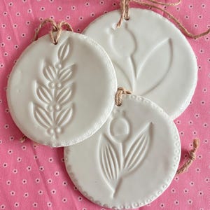 Può includere: Tre ornamenti rotondi in ceramica bianca con disegni botanici in rilievo, tra cui rametti e tulipani, appesi a spago su uno sfondo rosa floreale. Gli ornamenti hanno un aspetto rustico e fatto a mano.