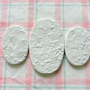 Puede incluir: Tres posavasos de cerámica blanca con diseños florales en relieve. Los posavasos son ovalados y redondos, con patrones detallados de hojas y flores. Están dispuestos sobre una tela a cuadros rosa y blanca.