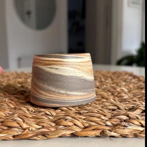 Peut inclure: Un petit gobelet en céramique fait à la main avec un motif marbré unique. Les couleurs comprennent des nuances de marron, de gris et de crème, créant une esthétique naturelle et terreuse. Le gobelet est posé sur un dessous de plat tressé, suggérant un thème rustique ou bohème.
