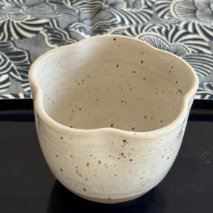 Puede incluir: Un cuenco de cerámica blanquecina moteada con un borde ondulado. El cuenco tiene un aspecto rústico y hecho a mano y se asienta sobre una superficie oscura. El fondo presenta una tela estampada en azul y blanco.