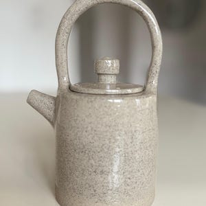 Può includere: Una teiera in ceramica grigia macchiata con un corpo arrotondato e un'unica maniglia. La teiera ha un coperchio con un piccolo pomo in cima.