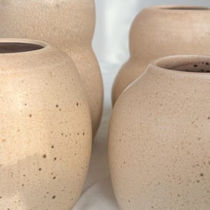 Pode incluir: Quatro vasos de cerâmica bege com superfícies salpicadas. Os vasos têm corpos arredondados e alturas variáveis, com uma paleta de cores neutras. Os vasos estão dispostos próximos uns dos outros, mostrando suas formas e texturas únicas.