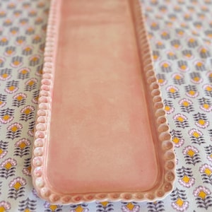 Puede incluir: Una bandeja rectangular de cerámica rosa claro con borde festoneado. La bandeja vacía descansa sobre una tela estampada con diseños florales en amarillo, rosa y gris.