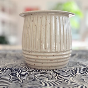 Könnte beinhalten: Eine gesprenkelte, cremefarbene Keramikvase mit vertikalen Rippendetails. Die Vase hat einen leicht ausgestellten Rand und eine zylindrische Form. Die neutrale Farbe und die strukturierte Oberfläche verleihen ihr ein rustikales, handwerkliches Aussehen. Die Vase steht auf einem gemusterten Stoff.