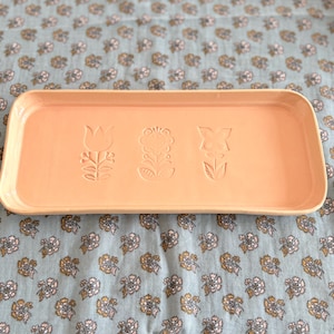 Puede incluir: Una bandeja rectangular de cerámica color melocotón con esquinas redondeadas. La bandeja presenta tres diseños florales en relieve: un tulipán, una flor estilizada y una flor de cuatro pétalos. La bandeja descansa sobre una tela azul con estampado floral.
