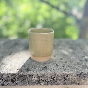 Op de afbeelding: Een handgemaakte keramische beker met een vierkante bovenkant en een lichtgroen glazuur. De bodem van de beker is naturel beige van kleur. De beker staat op een grijs stenen oppervlak met een vage groene achtergrond.