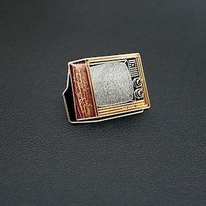 Può includere: Uno spillo in smalto dorato a forma di televisore vintage con un design in legno nero e marrone. Lo schermo mostra interferenze.