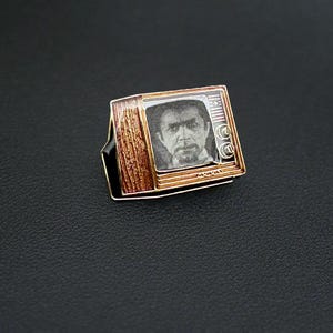 Könnte beinhalten: Ein Ring im Vintage-Stil mit einem Miniaturfernseher. Der Fernseher hat einen goldfarbenen Rahmen, einen Bildschirm mit einem Schwarz-Weiß-Bild eines Mannes und eine Seite in Holzoptik. Der Ring hat eine schwarze Basis.