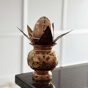 Op de afbeelding: Een koperkleurige decoratieve pot met ingewikkelde details, met groene en gouden accenten. De pot heeft een bolvormige basis en een deksel met een bladachtig ontwerp. De pot wordt waarschijnlijk gebruikt voor religieuze of ceremoniële doeleinden.