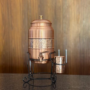 May include: A copper water dispenser with a hammered texture band, a spigot, and a lid. It sits on a black wrought iron stand with a matching cup holder and a copper cup. The dispenser is designed for serving water.